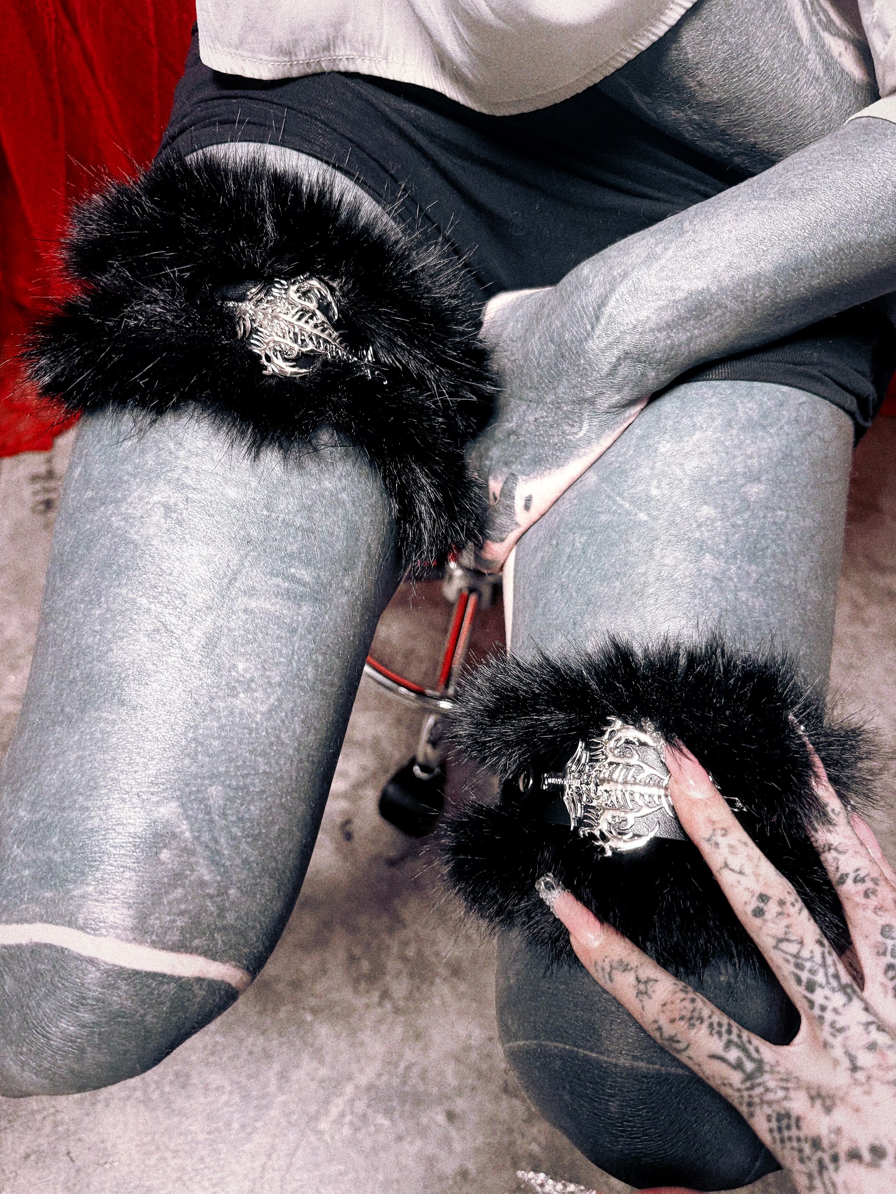 A close-up of a person seated on a chair wearing metallic silver thigh-high stockings with black furry cuffs and ornate silver medallions. The person's tattooed hand rests near the lower stocking, and a red fabric backdrop is visible in the upper left.