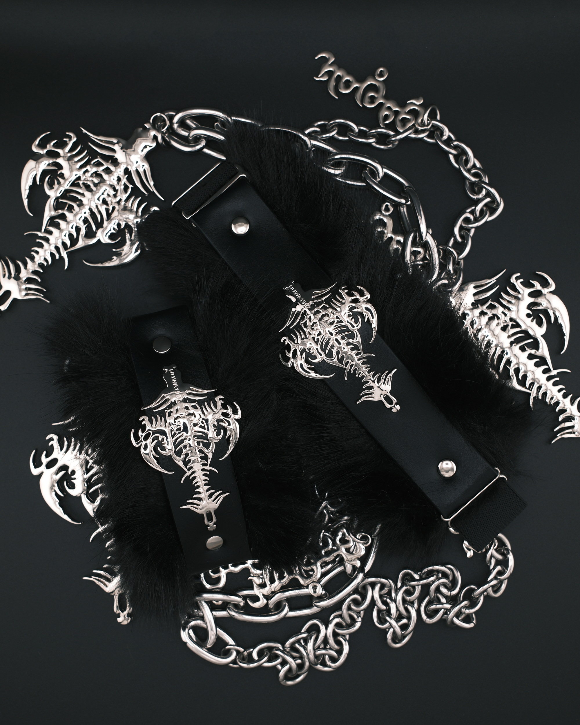 Black faux-fur cuffs with black leather straps and silver-toned metal hardware, decorated with gothic skeleton key and chain motifs, arranged on a dark background.