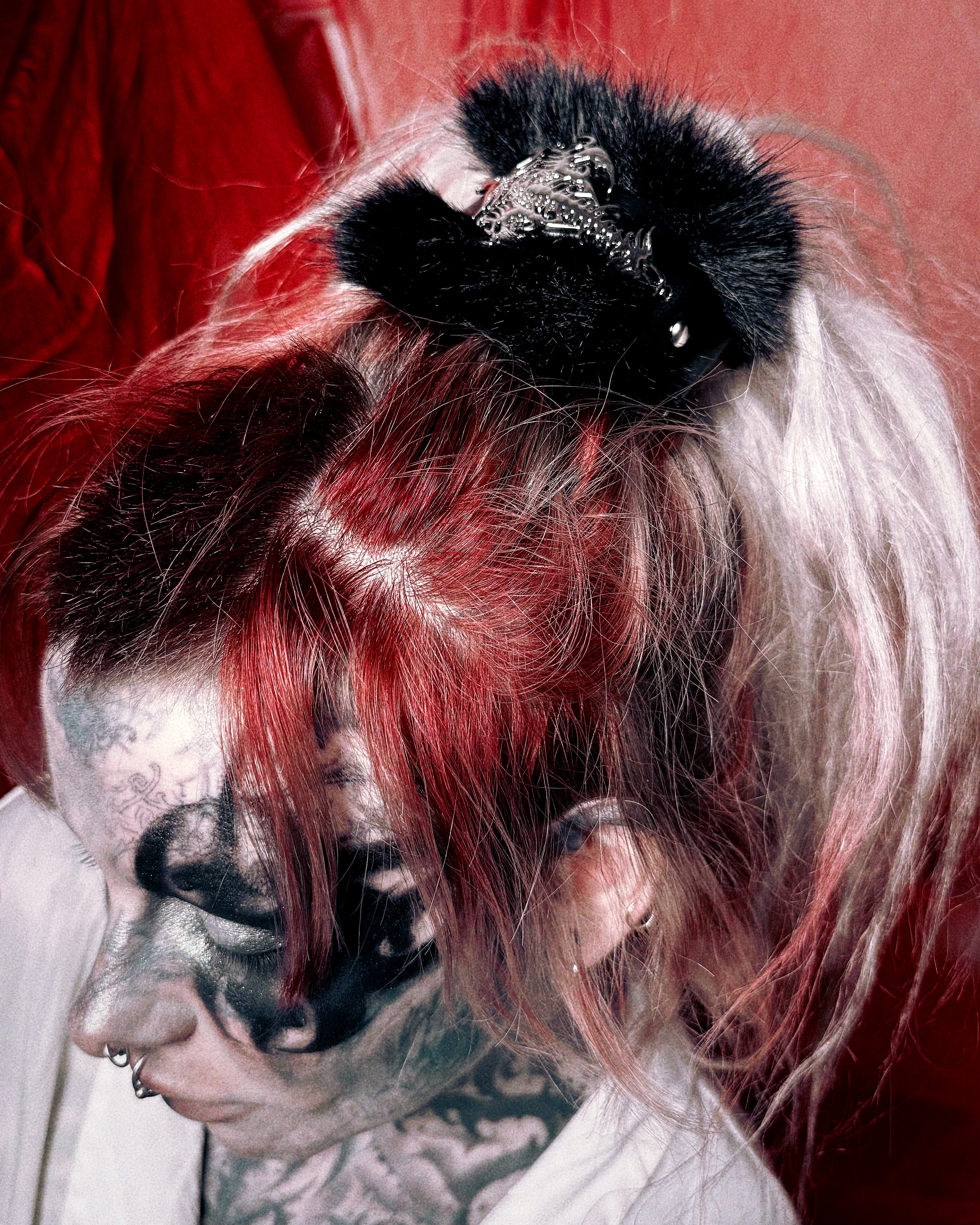 A close-up of a person with dramatic red, black, and white hair, wearing a black furry hair accessory with a silver decorative top. The person has face tattoos and gothic-style makeup, set against a dark red background.