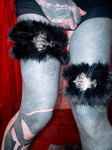 Black faux fur thigh-high stockings with silver metallic gothic-style embellishments, shown on a tattooed leg against a red background.