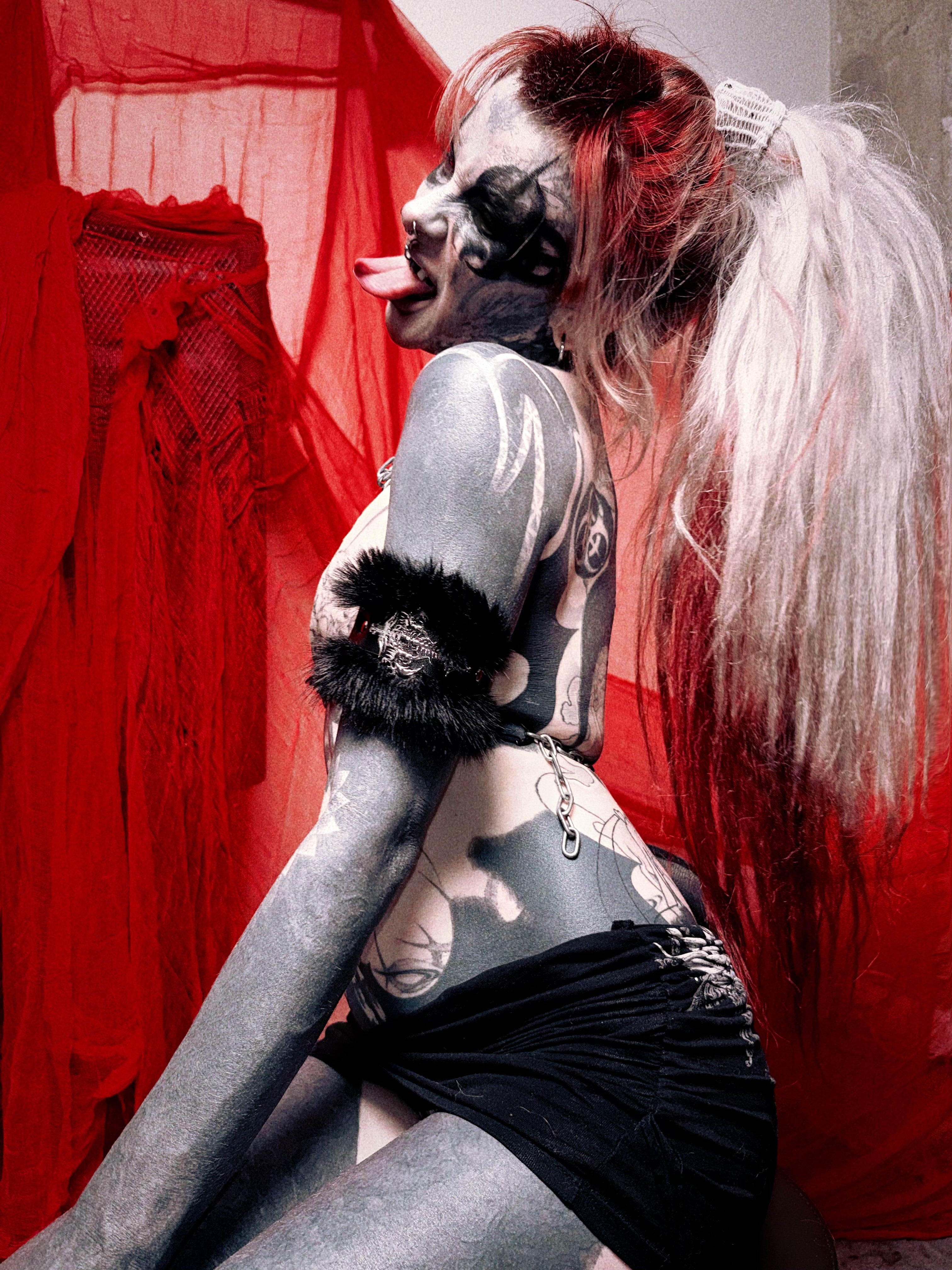 A tattooed model posing in a gothic style with pale face paint, a black ruffled armband, and a black ruffled headband, set against a background of red sheer fabric.