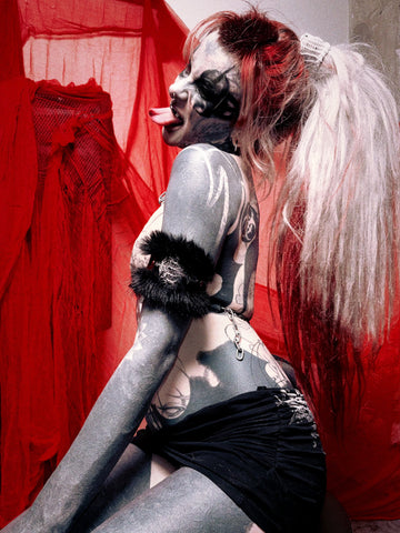 A tattooed model posing in a gothic style with pale face paint, a black ruffled armband, and a black ruffled headband, set against a background of red sheer fabric.