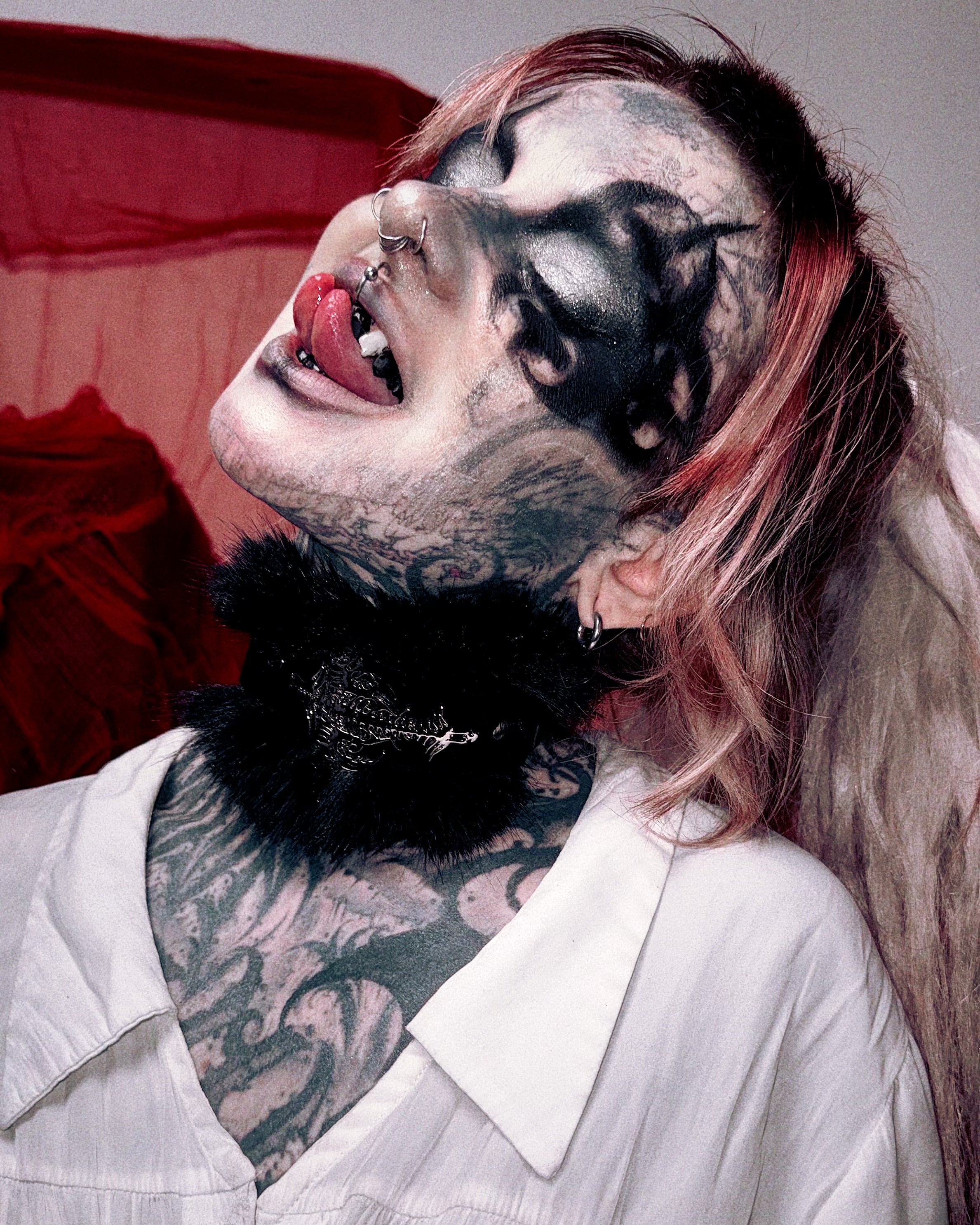 A close-up lifestyle shot of a person with gothic makeup, black feathered neckwear, and tattoos, wearing a white collared shirt against a dark red and gray background.