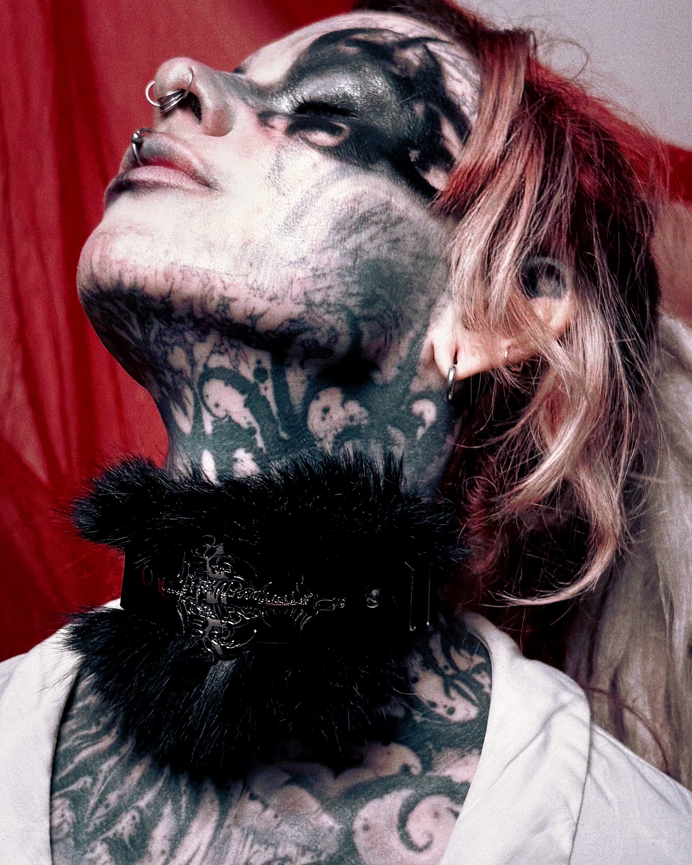 A close-up lifestyle shot of a person with extensive black and gray tattoos on their neck and chest, wearing a black furry choker with silver gothic filigree detailing. The background is a solid red curtain.