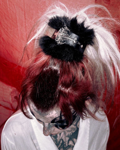 A close-up shot of a person with a messy high ponytail featuring a black faux-fur trim and a silver gothic filigree hair clip. The hair is dyed with dark red and platinum blonde streaks, and the person is wearing a white robe against a textured red background.