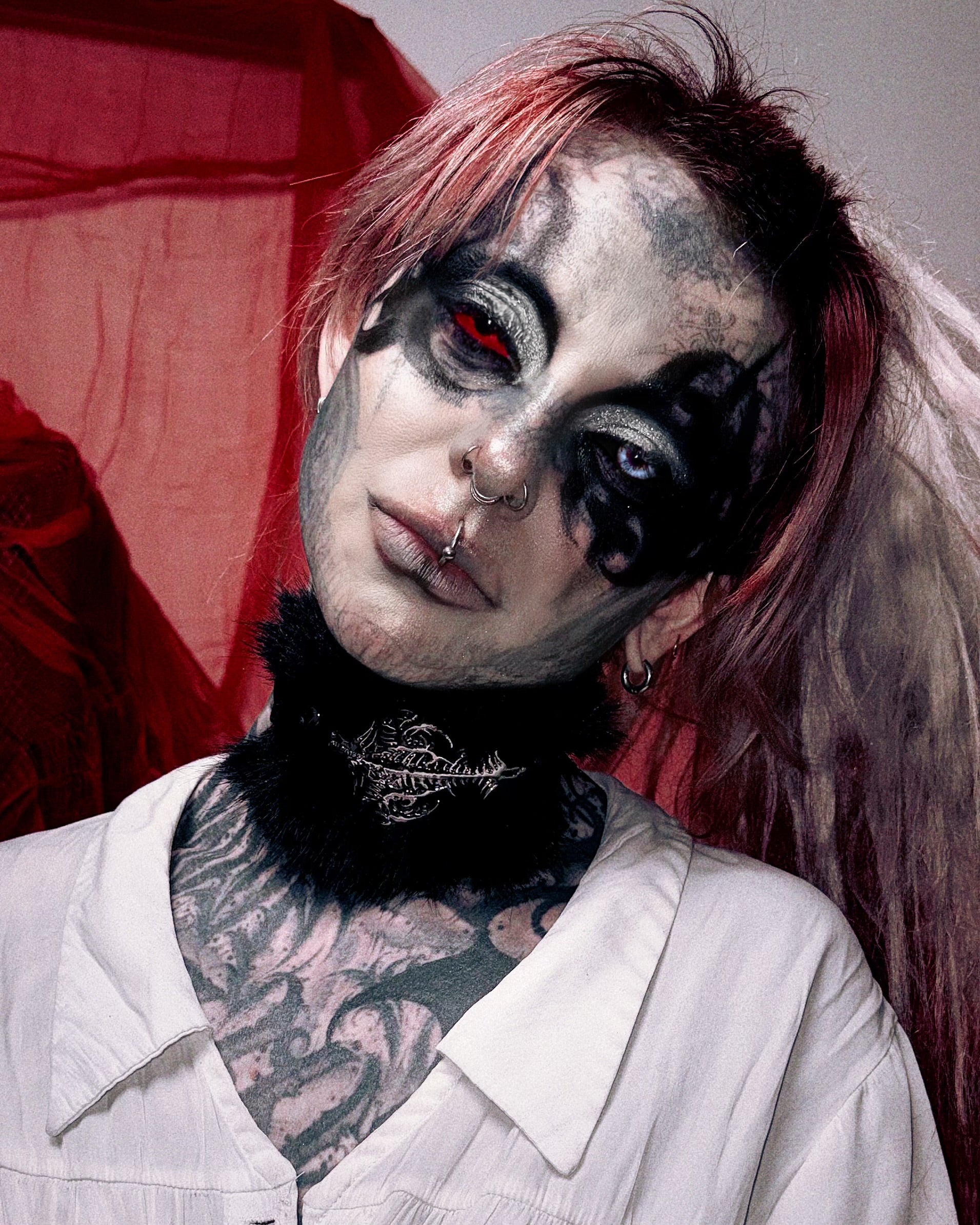 A close-up portrait of a person with dramatic gothic makeup, black tattoos, red hair, and a white collared shirt against a dark red and gray background.