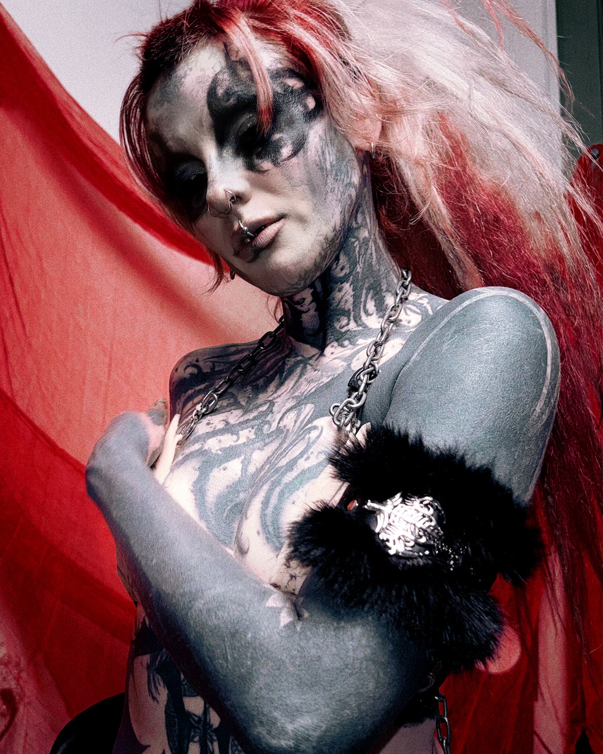A tattooed woman with pale makeup and red-and-pink hair poses against a red fabric backdrop, wearing a black furry armband with a silver rhinestone embellishment and a silver chain necklace.