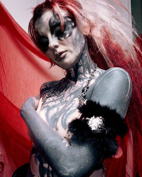 A tattooed woman with pale makeup and red-and-pink hair poses against a red fabric backdrop, wearing a black furry armband with a silver rhinestone embellishment and a silver chain necklace.