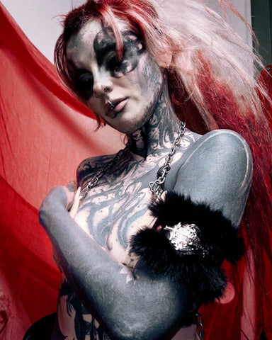 A tattooed woman with pale makeup and red-and-pink hair poses against a red fabric backdrop, wearing a black furry armband with a silver rhinestone embellishment and a silver chain necklace.